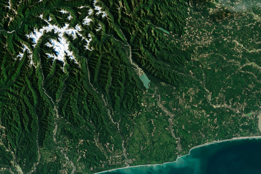 A satellite photo of the earth from above. The photo focuses on a piece of wooded coastline with some small lakes and a number of mountains covered in forest. There is a small amount of snow covering the tops of the peaks.